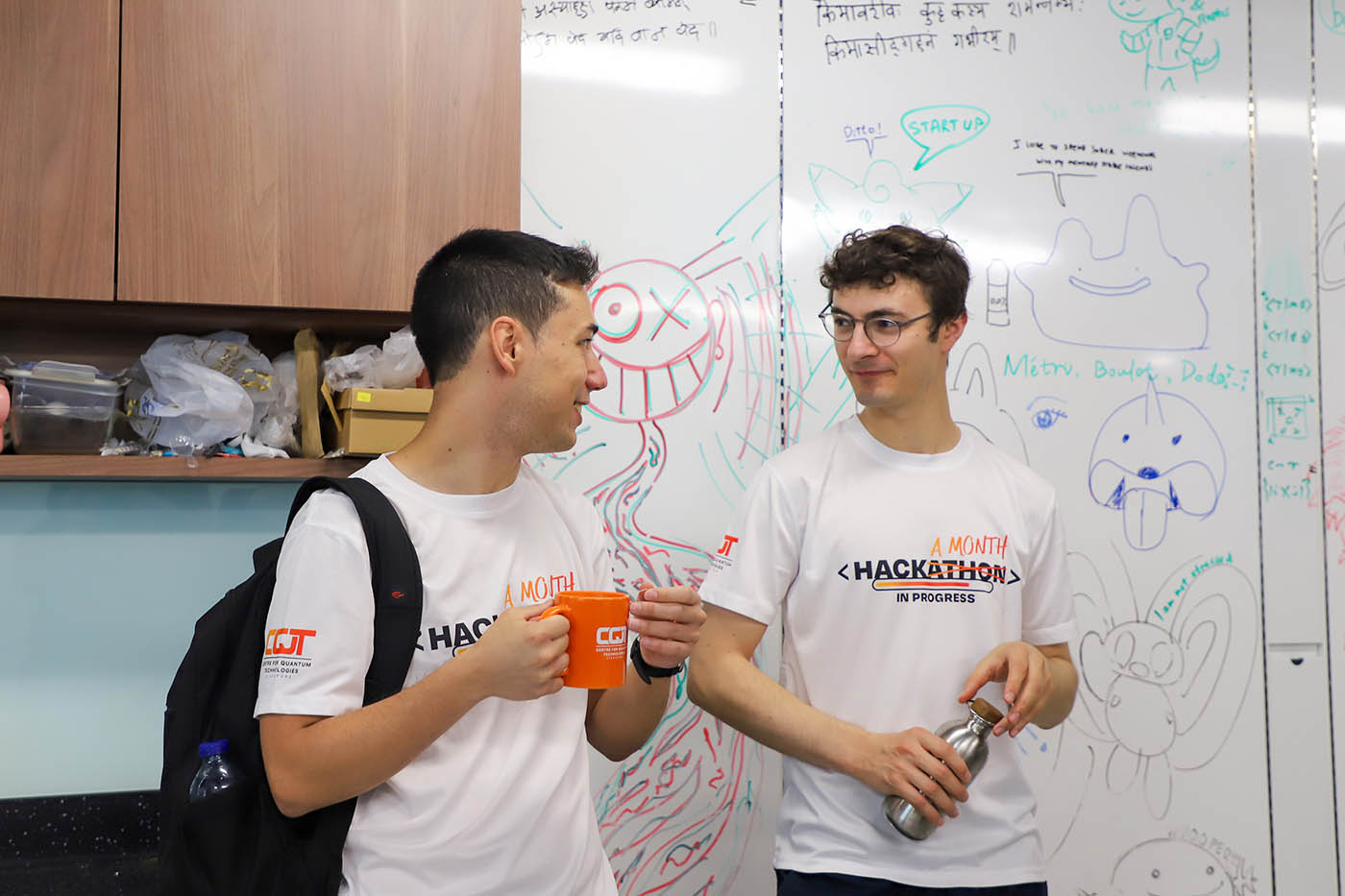Two men in conversation in front of a whiteboard covered in cartoon drawings. Both where t-shirts saying Hackamonth in progress.