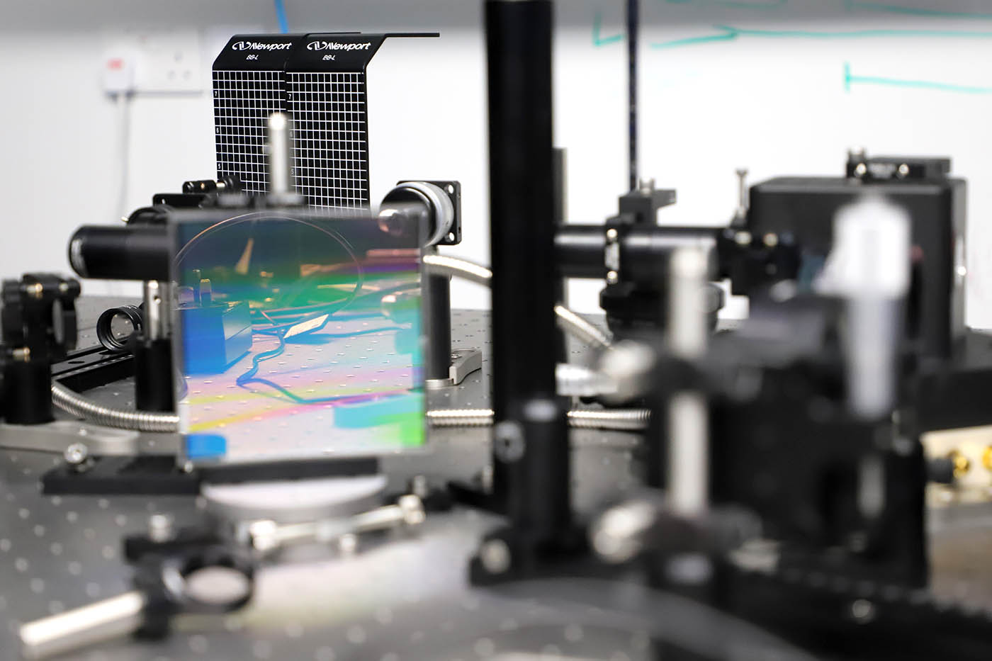 A close-up look at optical components in the experiment. The focus is on a square sheet of diffraction grating that has a rainbow sheen.
